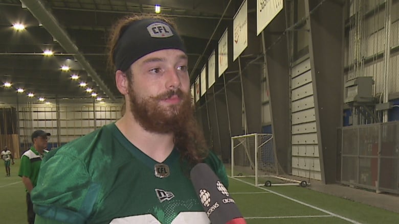 A white man with long brown hair and a thick, scruffy brown beard, is wearing a green football jersey and a black headband with the CFL logo on the front. He is standing on an indoor turf field.