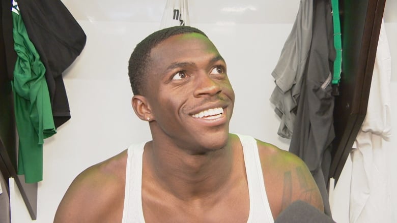 A clean-shaven Black man, with short dark hair, is smiling. He is wearing a white tanktop, while sitting at a locker.