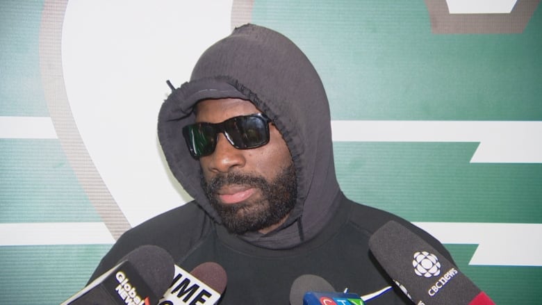 A Black man with a thick dark beard is wearing a black hoodie with the hood up and sunglasses. He is standing in front of four microphones.
