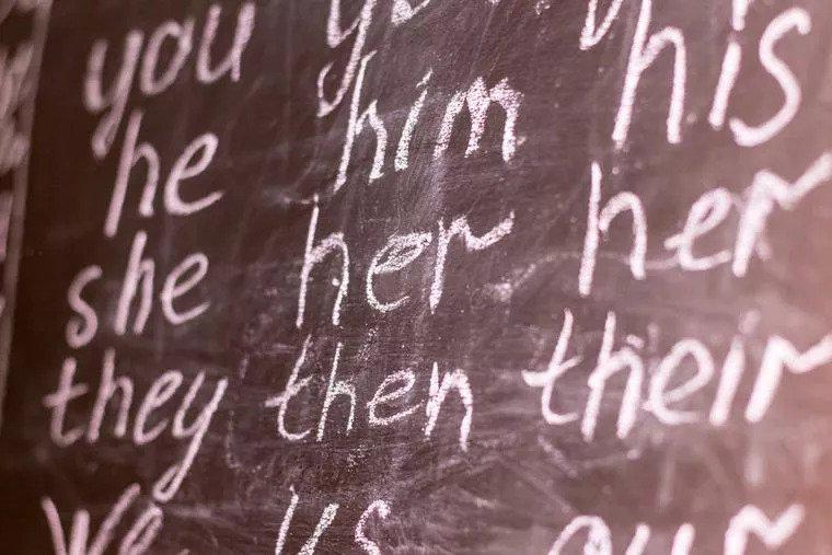 Gender pronouns written out in cursive on a chalkboard inside a classroom.