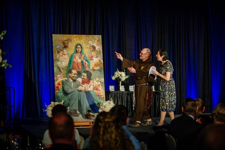 Above, Father Joseph Christensen unveils a commissioned Marian-themed painting featuring St. Gianna Molla and her husband, Pietro, during the ‘Canticle of Praise’ event honoring 20 years of the Saint Gianna & Pietro Molla Maternity Home in Grand Forks, North Dakota, on April 16. Here, he points out to Gianna Emmanuela Molla, the youngest child of St. Gianna, that the artist included a likeness of her sister Mariolina, who died at age 6. The home hopes to soon be able to procure the rights to sell the image of the Mollas on prayer cards.
