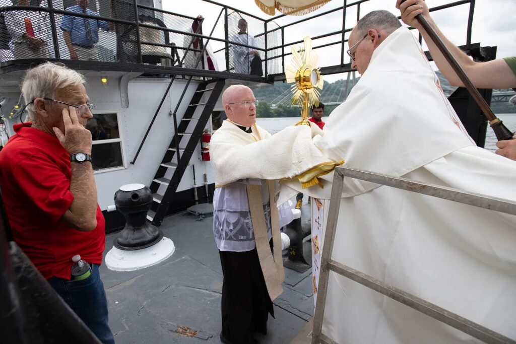 Upon arrival, Bishop Lohse hands Jesus Christ in the Blessed Sacrament to Fr. Roger Landry aboard the vessel ‘Sewickley.’