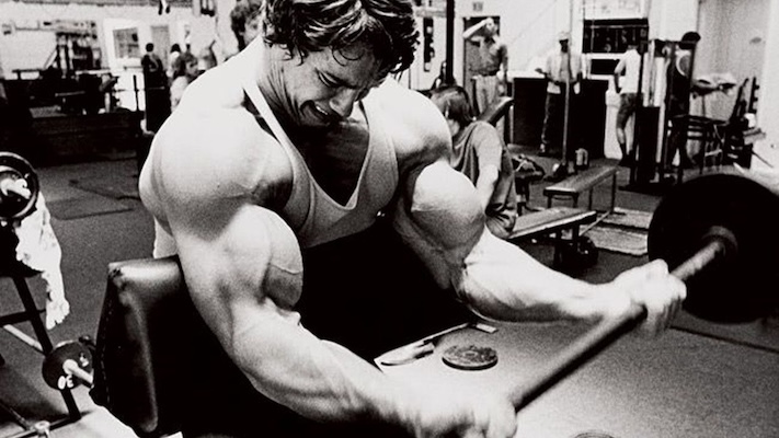 A person in a gym intensely performs a bicep curl with a barbell on a preacher bench. Other gym-goers and equipment are visible in the background.