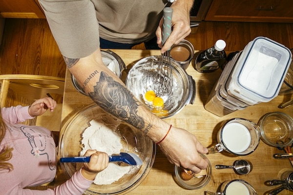 Overhead view of two people mixing Lazy Weekend Pancakes ingredients in bowls on a wooden kitchen table, surrounded by containers, measuring cups, and milk.
