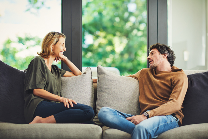 Two people sit on a sofa facing each other, engaged in conversation during a Marriage Checkup, with large windows and greenery visible in the background.