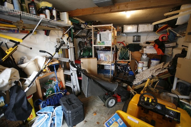 A cluttered garage with shelves full of boxes, tools, paint cans, and various stored items shows classic garage clutter. A lawnmower and a yellow toy vehicle are on the floor—perfect candidates for decluttering tips or things to get rid of.